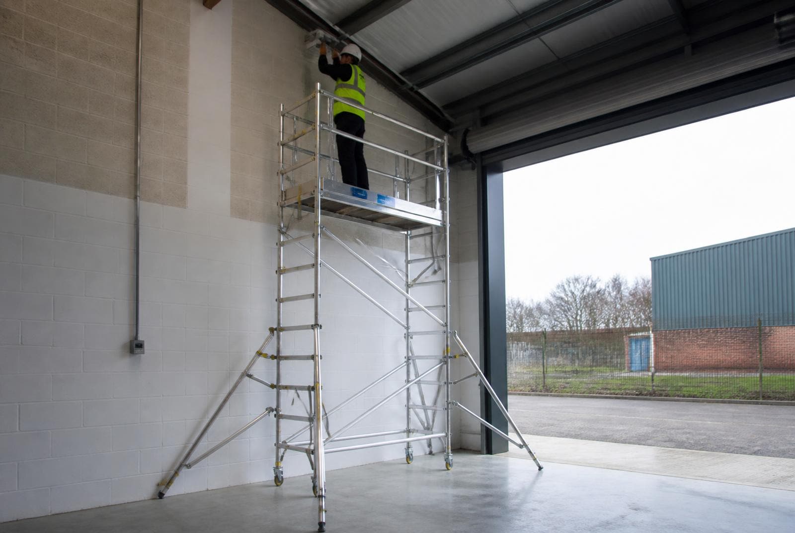 Aluminium mobile access tower with stabilisers and guard rails erected inside a UK commercial unit, with a worker on the platform.