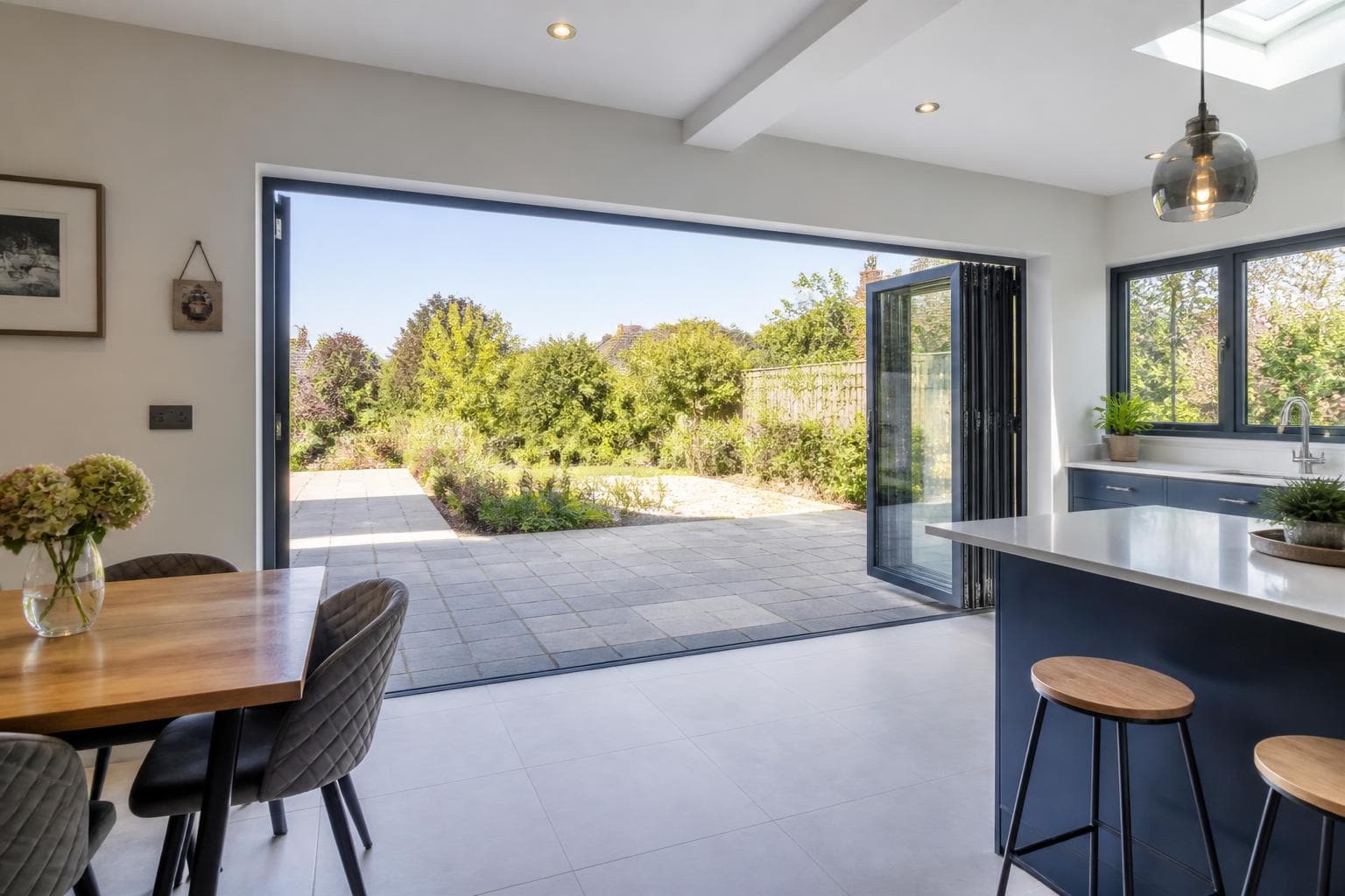Grey aluminium bi-fold doors folded fully open between a bright UK kitchen diner and a paved patio.
