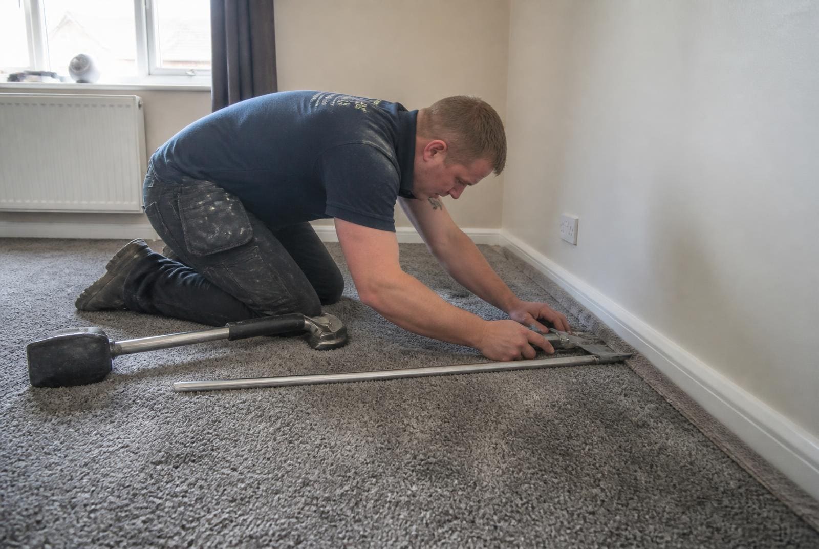 Carpet fitter using a knee kicker to stretch and trim new grey carpet against gripper rods in a UK bedroom.