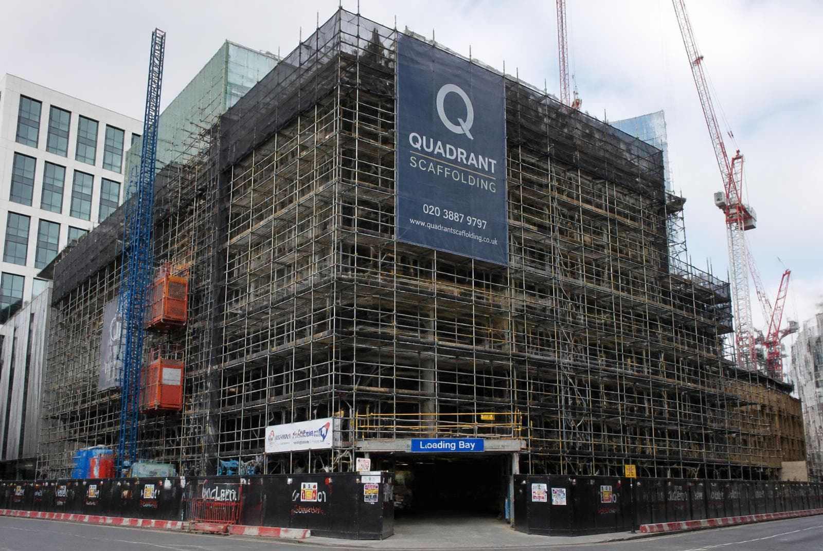 Large commercial scaffold with debris netting, hoist and loading bay wrapping a multi-storey UK city centre building.