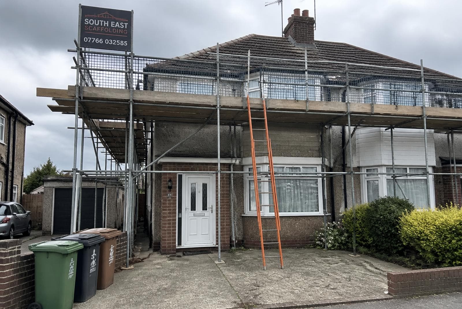 Domestic scaffolding erected around a UK semi-detached house for a re-roof, with edge protection, ladder access bay and a company sign board.
