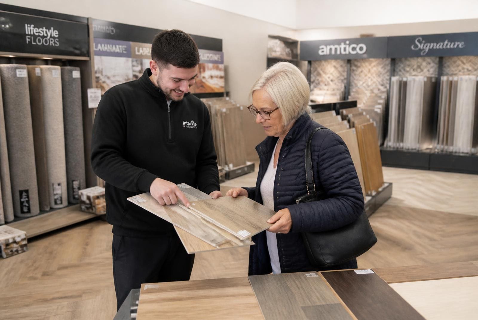 Flooring showroom staff member showing a customer LVT and laminate samples in front of racks of carpet rolls.