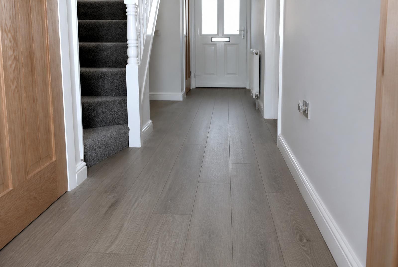 Grey plank laminate flooring newly fitted in a UK hallway with white skirting, scotia beading, stairs and a front door in view.