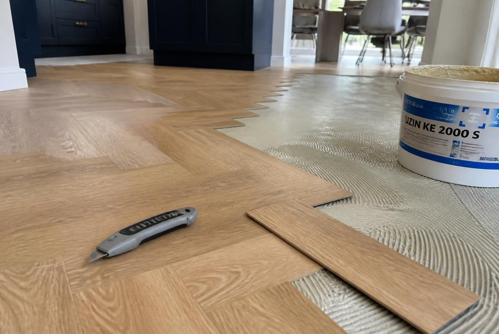 Luxury vinyl tile herringbone flooring being laid over adhesive in a modern UK kitchen diner with a trimming knife on the floor.