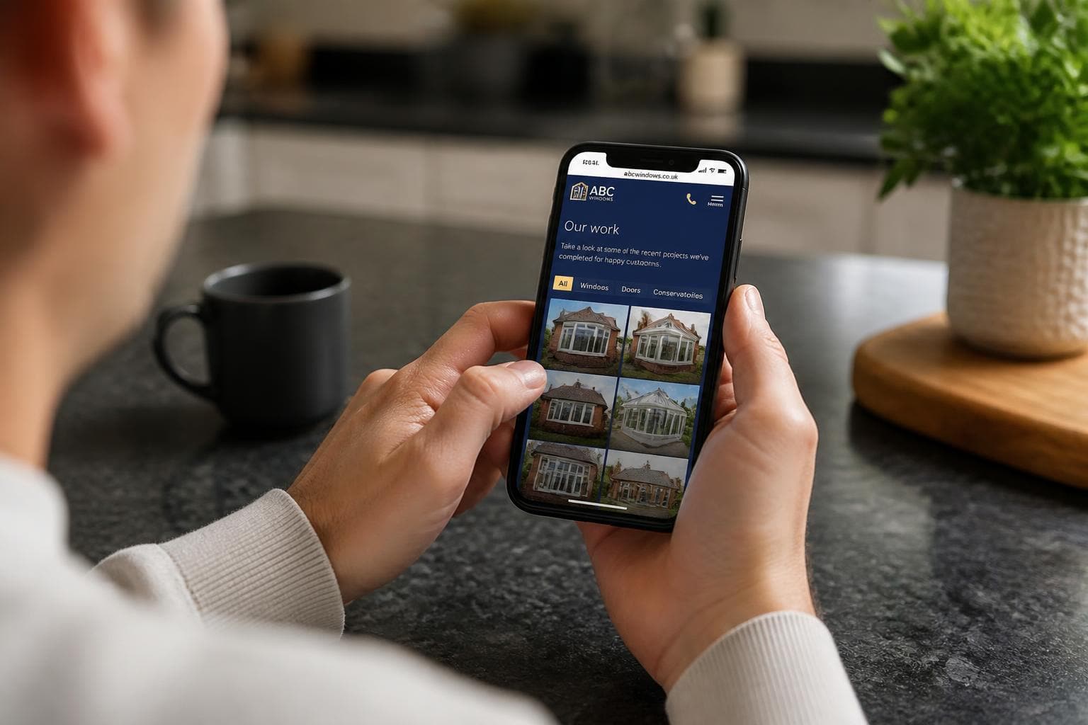 Homeowner viewing a double glazing company's project gallery on a smartphone at a kitchen worktop.