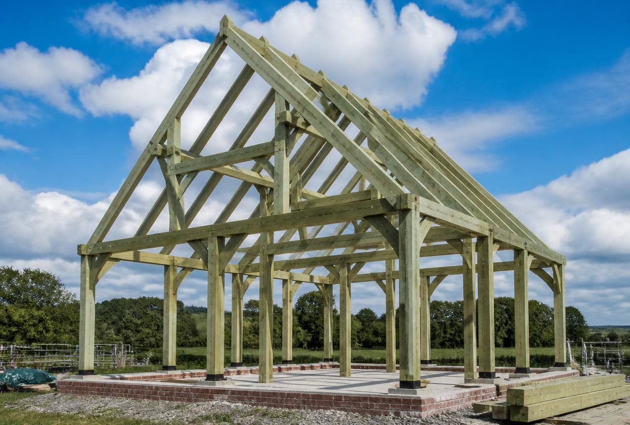 Green oak timber frame structure with mortise and tenon joints standing on a brick plinth in a rural UK field.