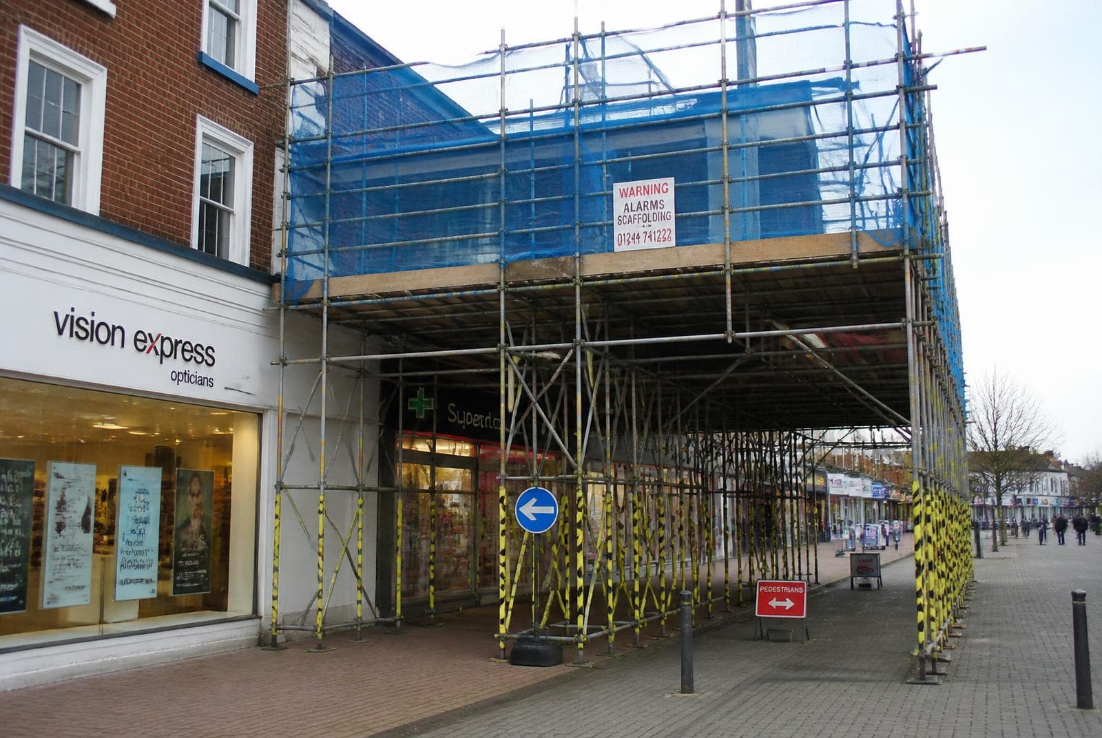 Pavement gantry and scaffold over a UK high street shop frontage with debris netting, warning signage and a protected pedestrian walkway.