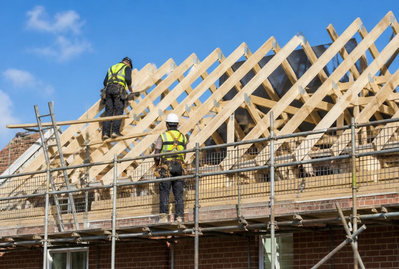 Two roof carpenters in high-visibility clothing fixing timber trusses and rafters on a UK house roof against a blue sky.
