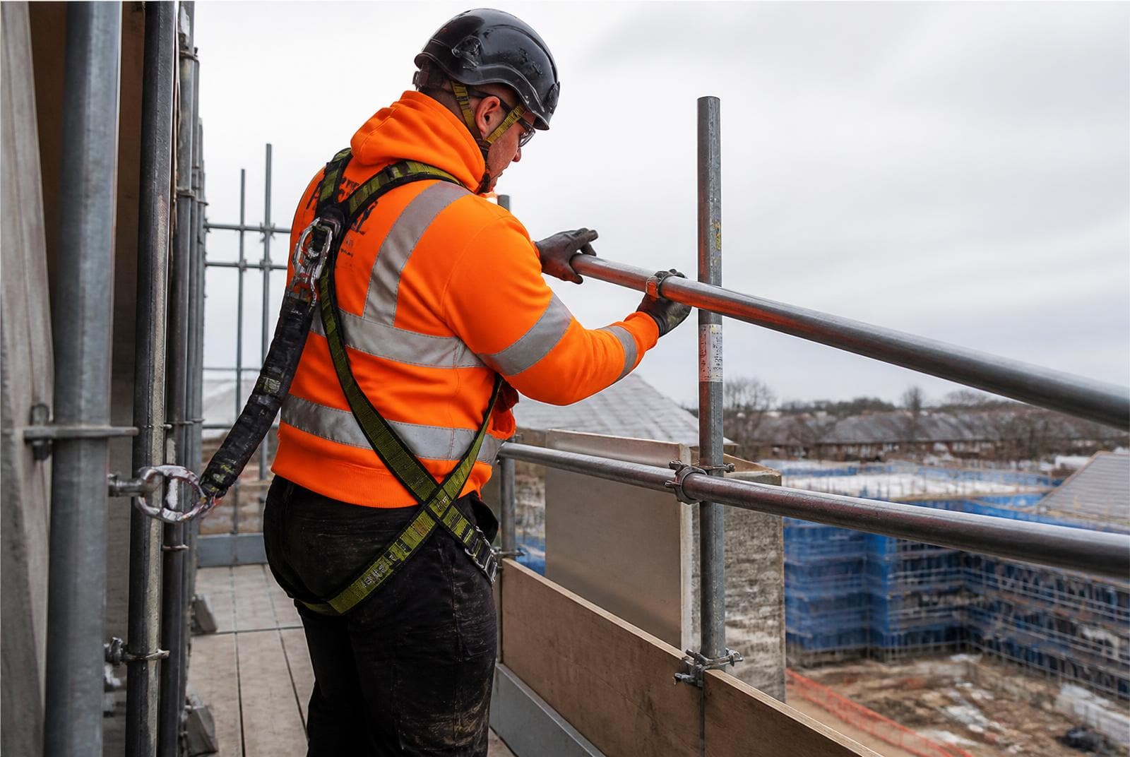 Scaffolder wearing a full body harness clipped on while fitting a guard rail at height on a scaffold lift above a construction site.