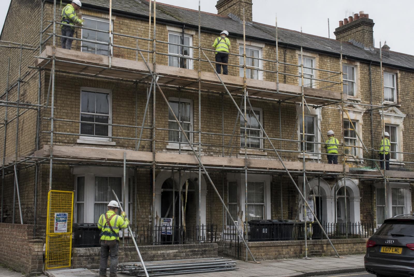 UK scaffolders in hi-vis and harnesses erecting a tube and fitting scaffold across the front of a Victorian terraced house.
