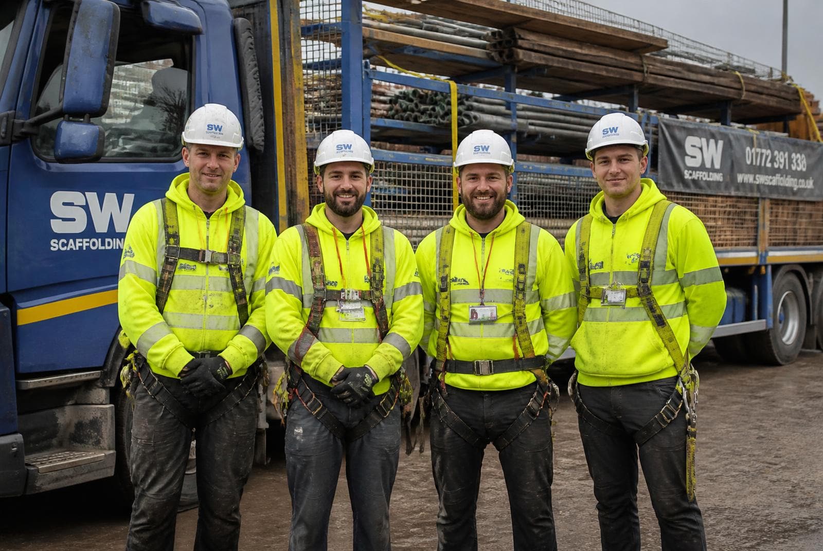 Team of four UK scaffolders in branded hi-vis, hard hats and harnesses standing in front of a loaded scaffolding lorry at their depot.