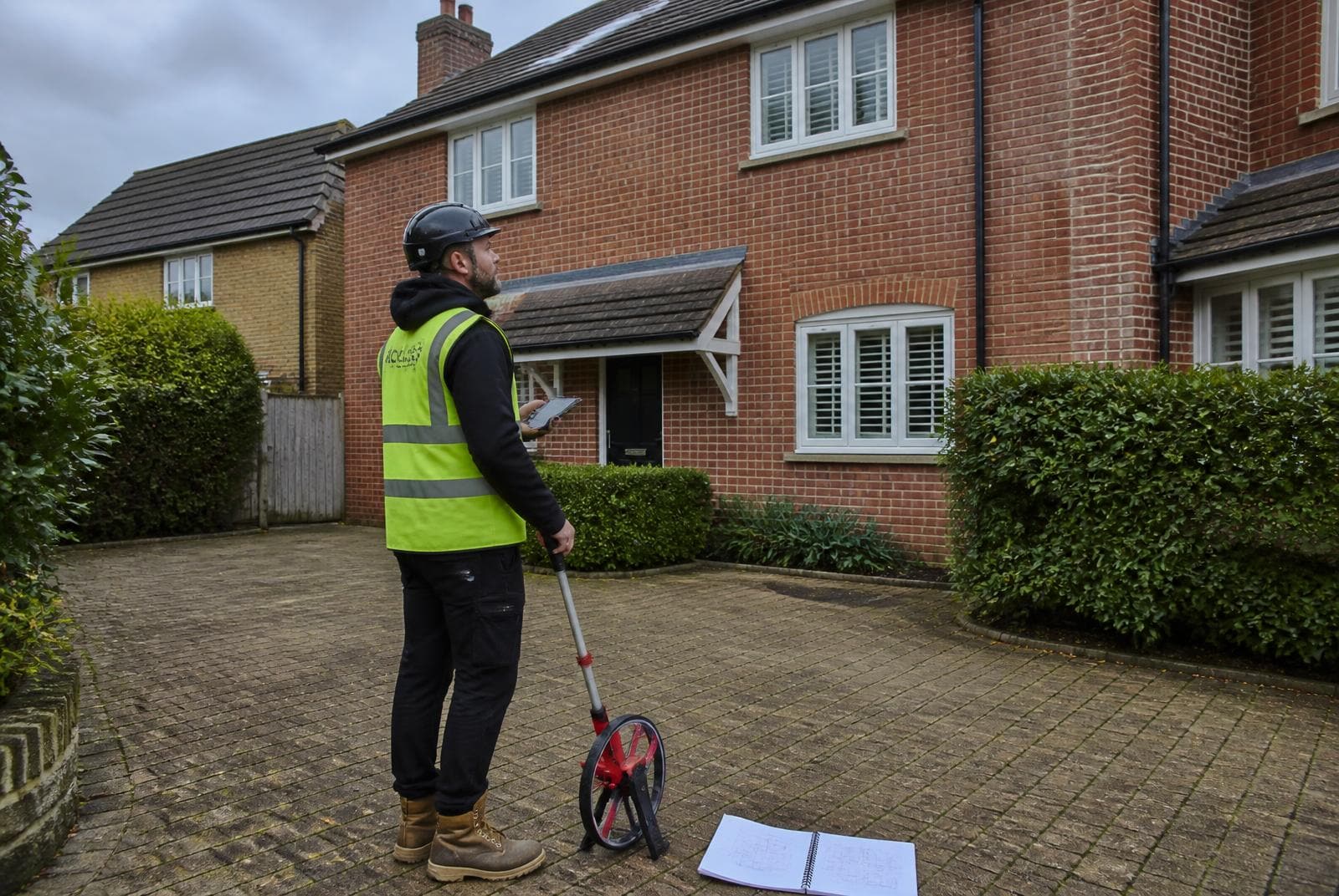 Scaffolding supervisor with a tablet and measuring wheel carrying out a site survey outside a UK house before erecting scaffold.